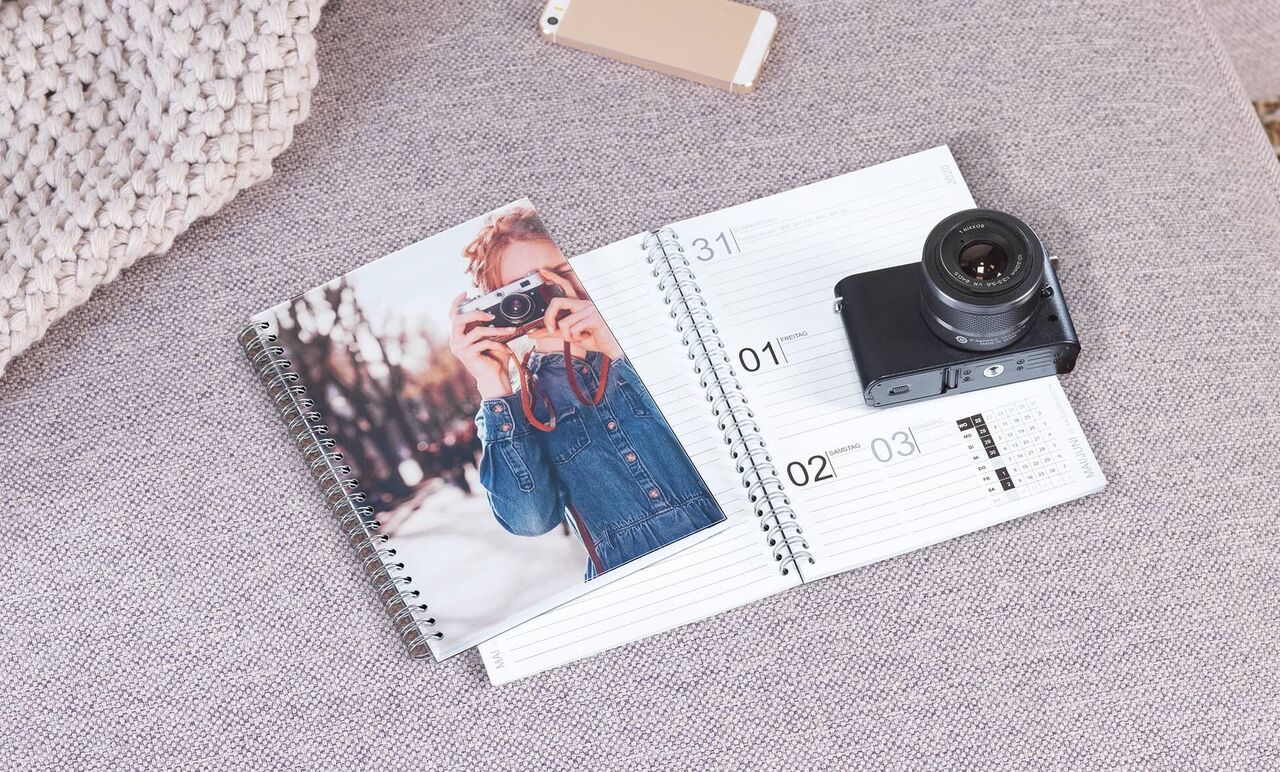 En fotokalender ligger på en grå sofa og viser et billede af en smilende fotograf. Ved siden af er et kamera og en mobil.