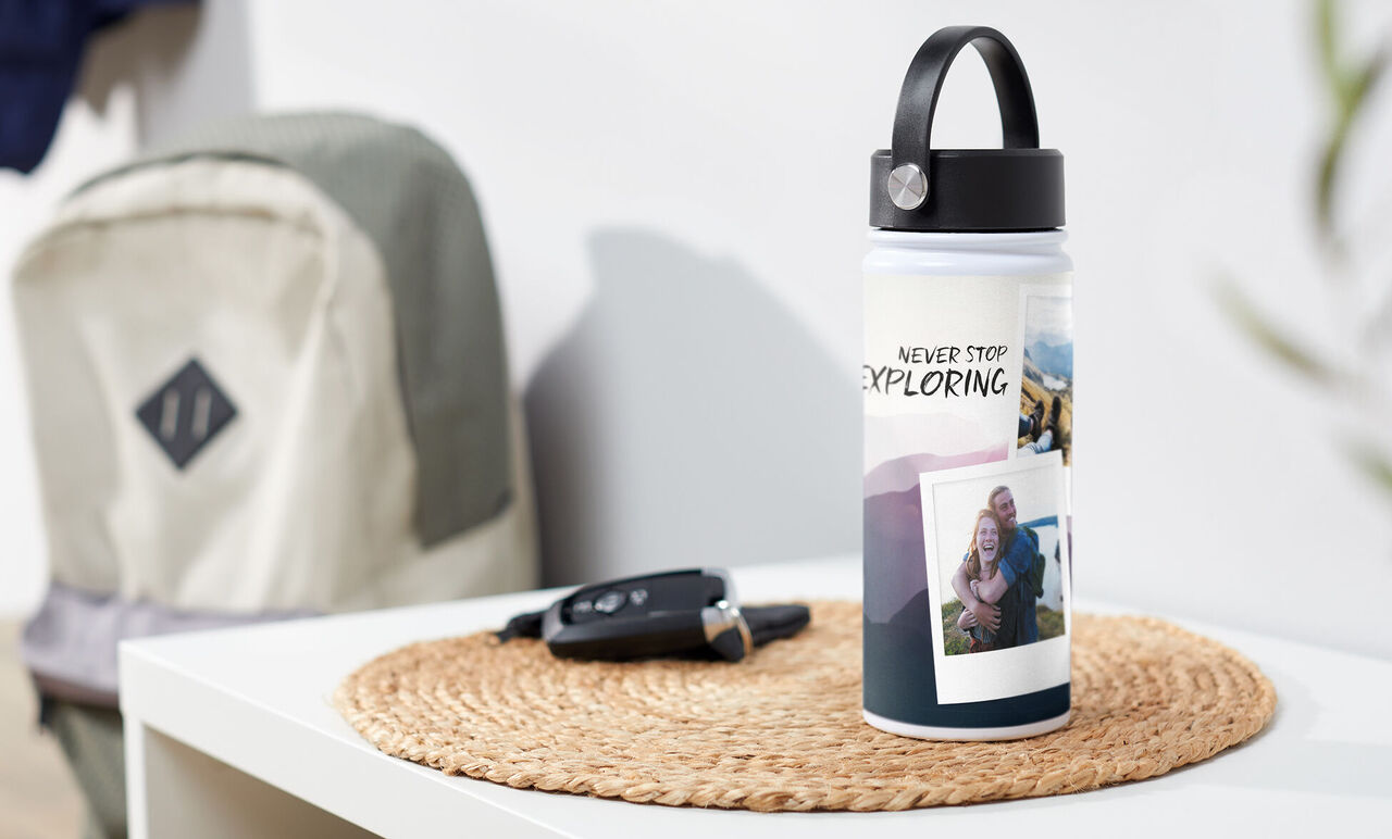Vandflaske med teksten 'Never Stop Exploring' og fotos på et bord. I baggrunden en rygsæk og bilnøgle.