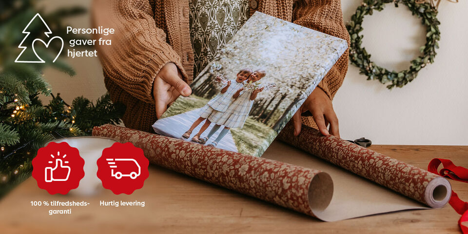 Et barn og en pige holder en fotobog med et billede af sig selv, mens de pakker gaver. Bag dem er julepynt.