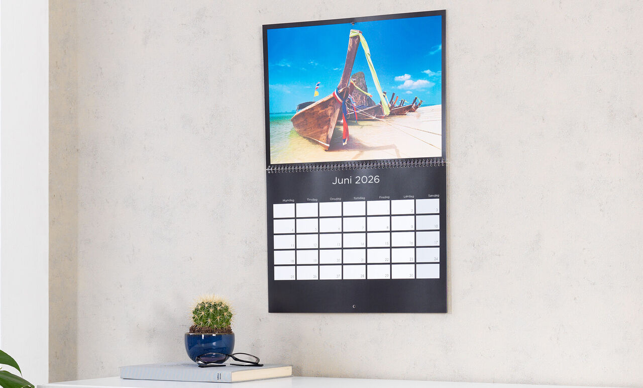 En vægkalender viser et billede af en båd på stranden, juni 2025.