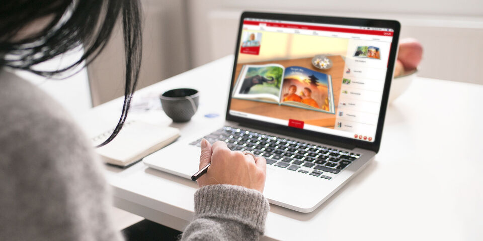 En person sidder ved et bord og ser på en bærbar, der viser CEWE FOTOBOG-hjemmesiden.