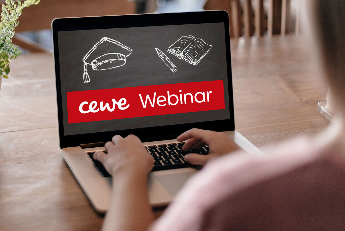 En person sidder foran en laptop og deltager i et CEWE-webinar. Skærmen viser en akademisk hat, en notesbog og en pen.