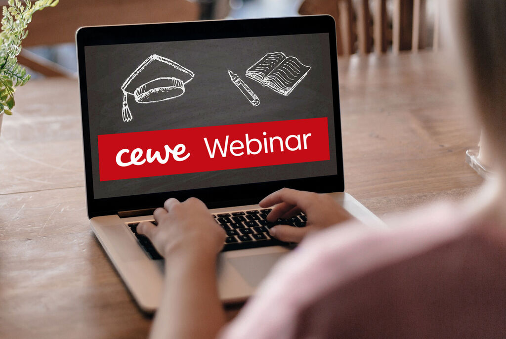 En person sidder foran en laptop og deltager i et CEWE-webinar. Skærmen viser en akademisk hat, en notesbog og en pen.