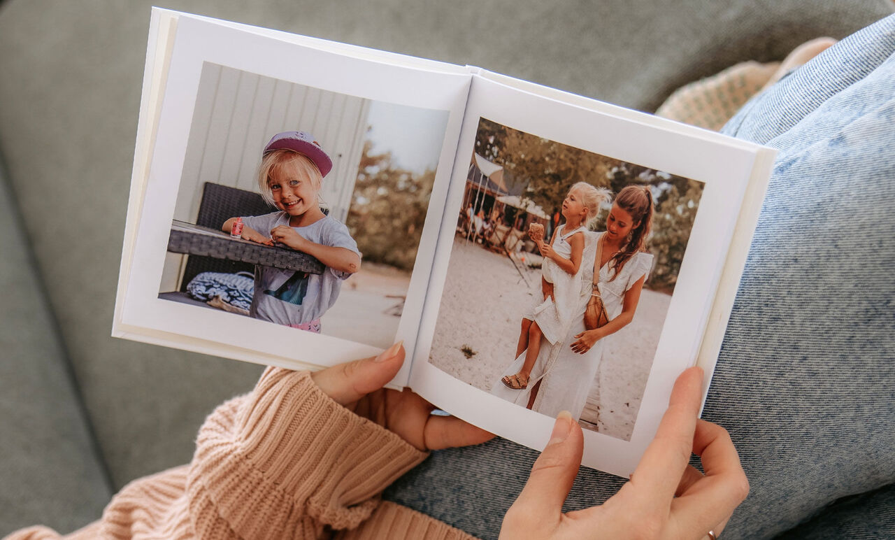 En person holder en åben fotobog med to billeder. Det første billede viser et barn ved et bord, det andet billede viser en kvinde med et barn.