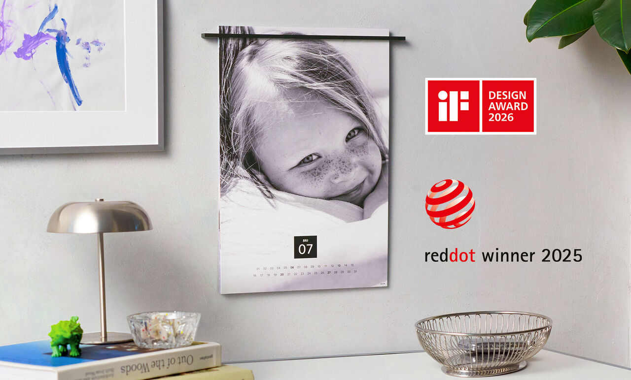 Vægkalender med et smilende barn og en smuk baggrund. Kalenderen viser juli måned og har red dot design award logo.