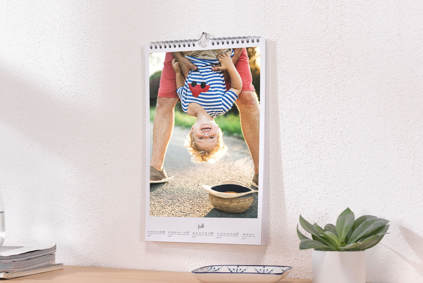 Vægkalender med et glad barn, der hænger med hovedet nedad og smiler, med en grøn græsplæne i baggrunden.