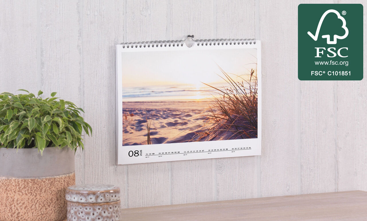 En smuk vægkalender med en solnedgang over en strand i august. En grøn plante står ved siden af.