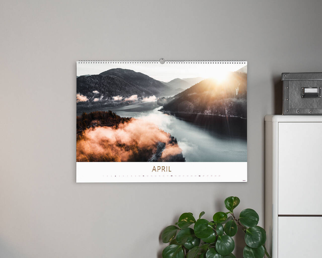Vidunderlig fotokalender med landskabsbillede til april. Kalenderen viser solopgang over en tåget sø, omgivet af grønne bjerge.