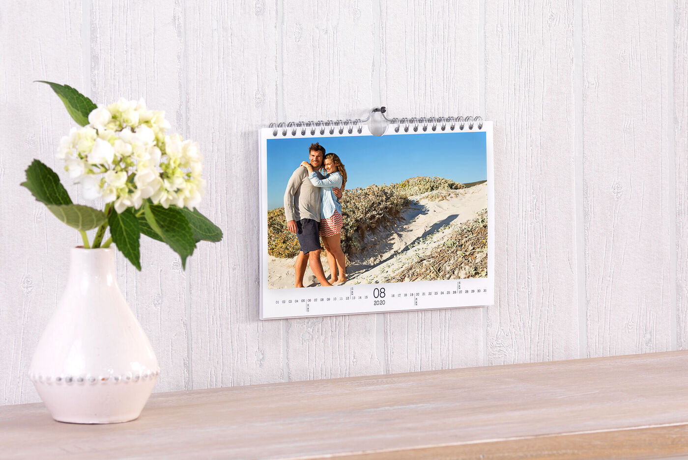 En kalender hænger på væggen. Kalenderbilledet viser et par, der krammer med en sandstrand i baggrunden.