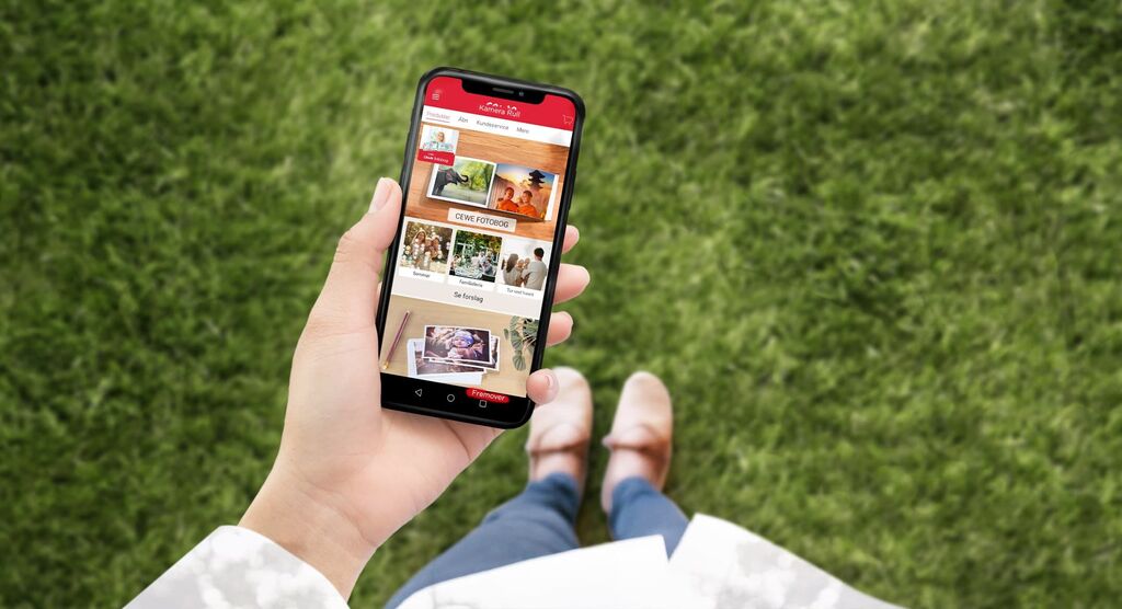 Hånd holder smartphone med åbnet CEWE-app på grønt græs
