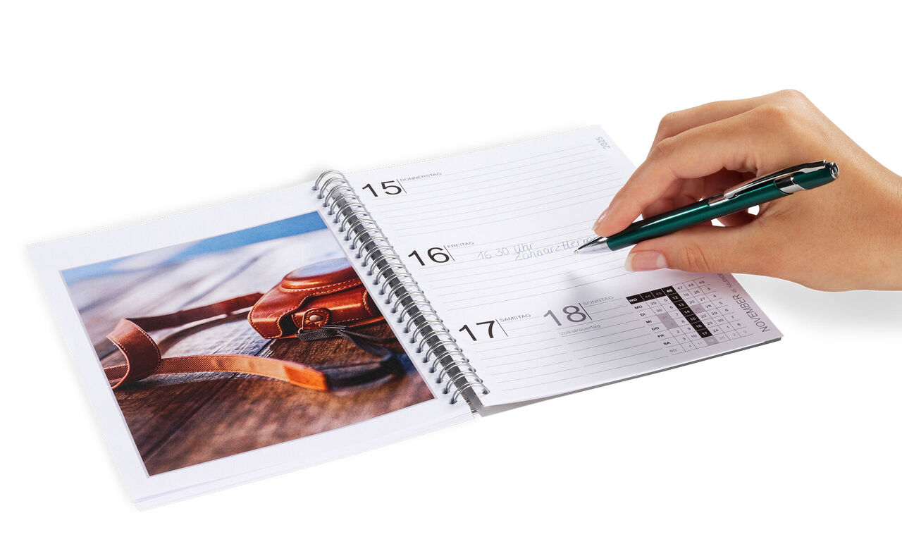 En hånd skriver i en kalender, ved siden af et foto med solbriller på et bord.