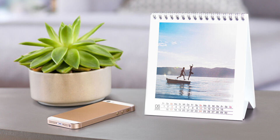 En lille bordkalender med et foto af to personer, der står på en båd og nyder vandsport. I baggrunden ses bløde bølger og en smuk natur.