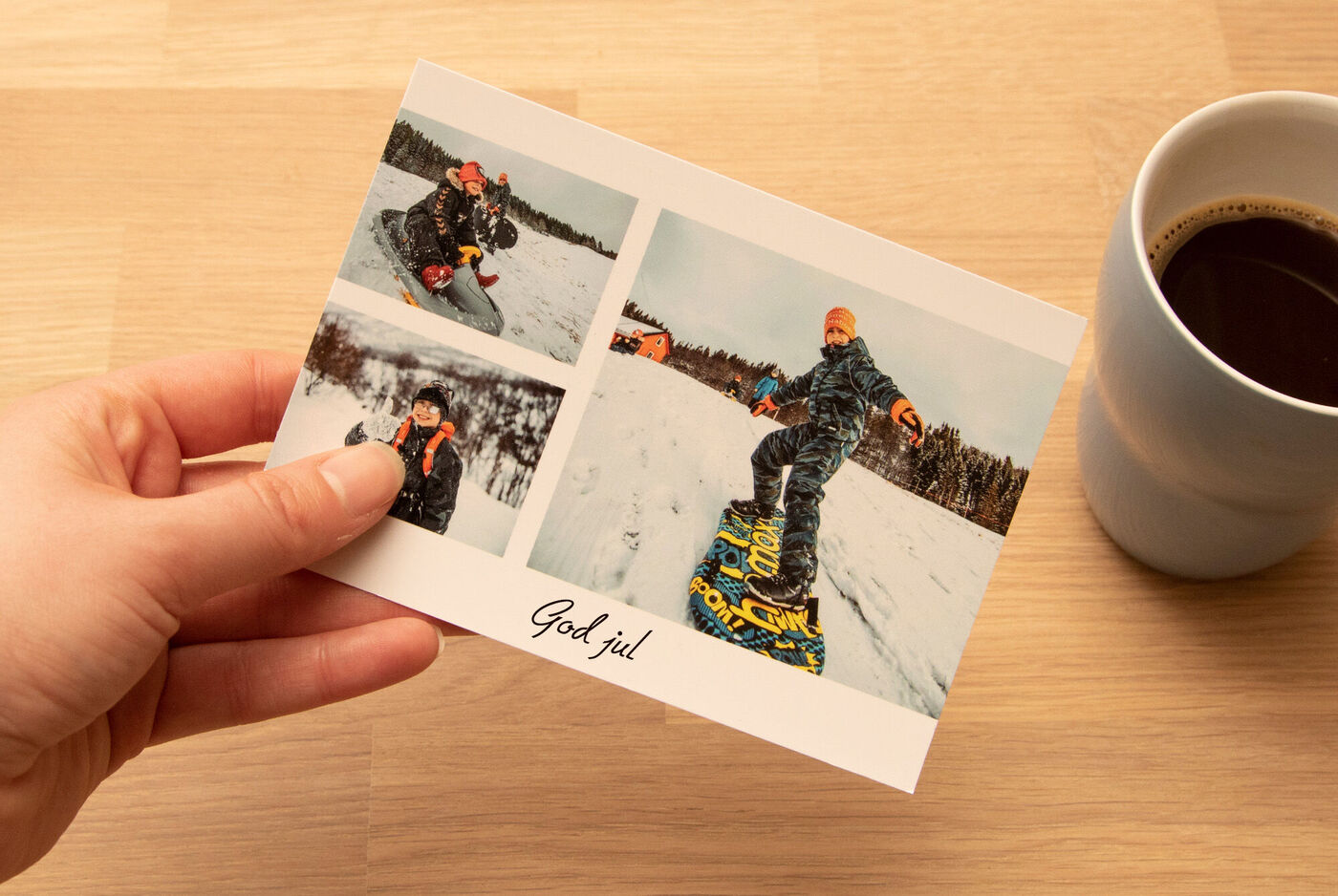En person holder et julekort med billeder af en snowboarder. På kortet står "God jul". Ved siden af kortet står en kop kaffe.