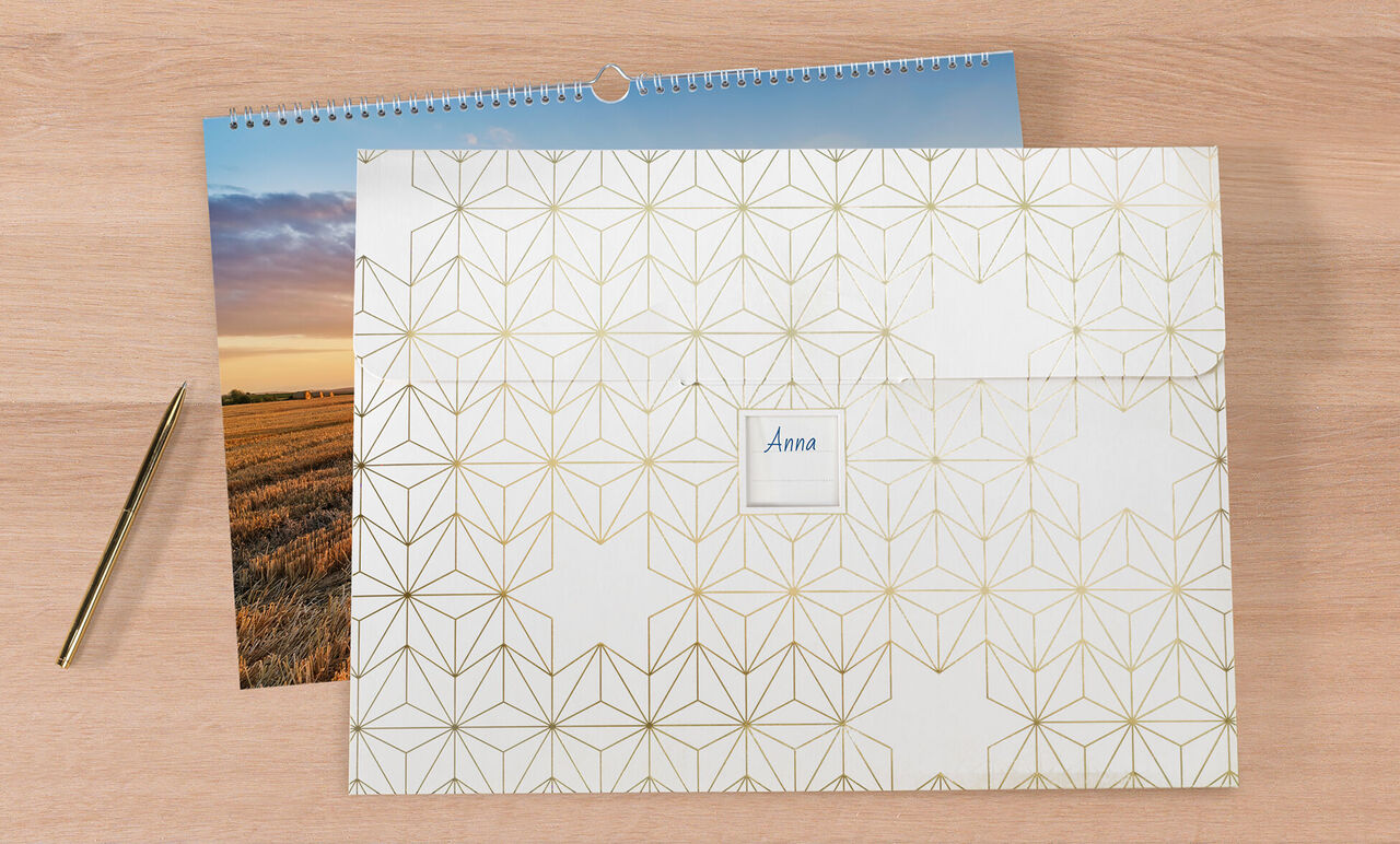 En lukket CEWE fotokalender på en træoverflade, ved siden af et foto og en guldpen.