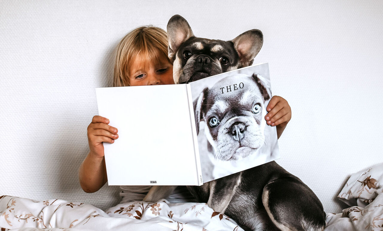 Et barn holder en bog med titlen 'THEO', mens en hund sidder ved siden af og ser på.