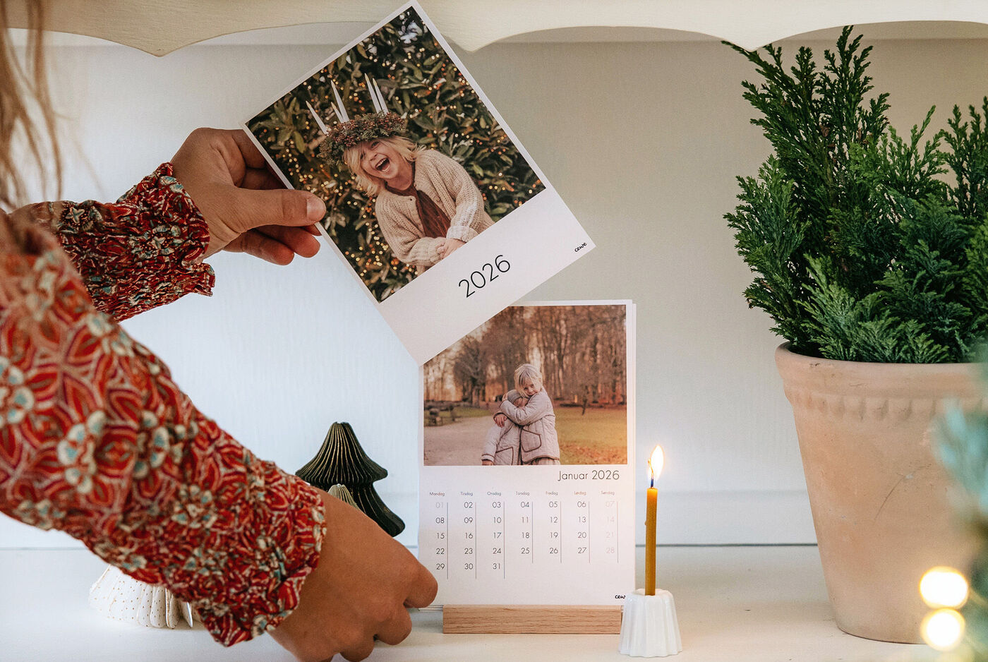 En person holder en fotograferet kalender med billeder og årstal 2025 på et bord. Et lys brænder ved siden af en juletræ.