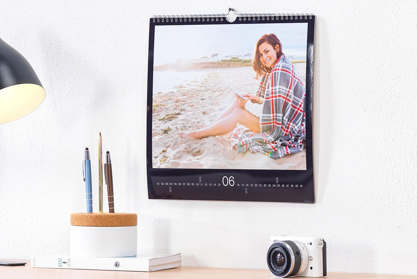 En CEWE fotokalender med et snapshot af en person, der sidder på stranden og læser en bog, lænet mod et bord med blyanter og et kamera.