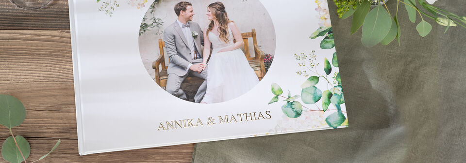 En smuk CEWE fotobog med titlen Annika & Matthias på et træt bord omgivet af grønne blade.