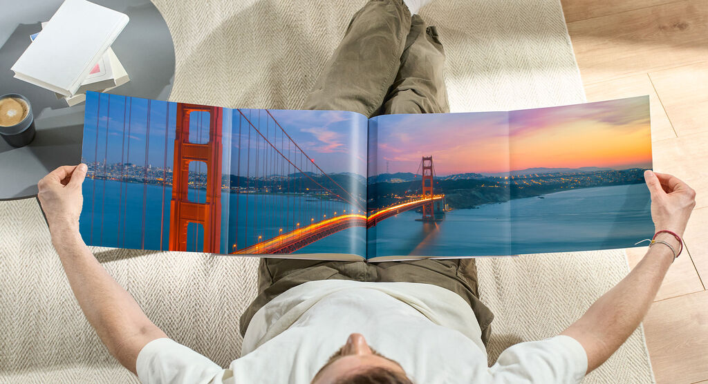 En person sidder behageligt på gulvet og ser på en åbnet fotobog med et smukt billede af Golden Gate Bridge ved solnedgang.