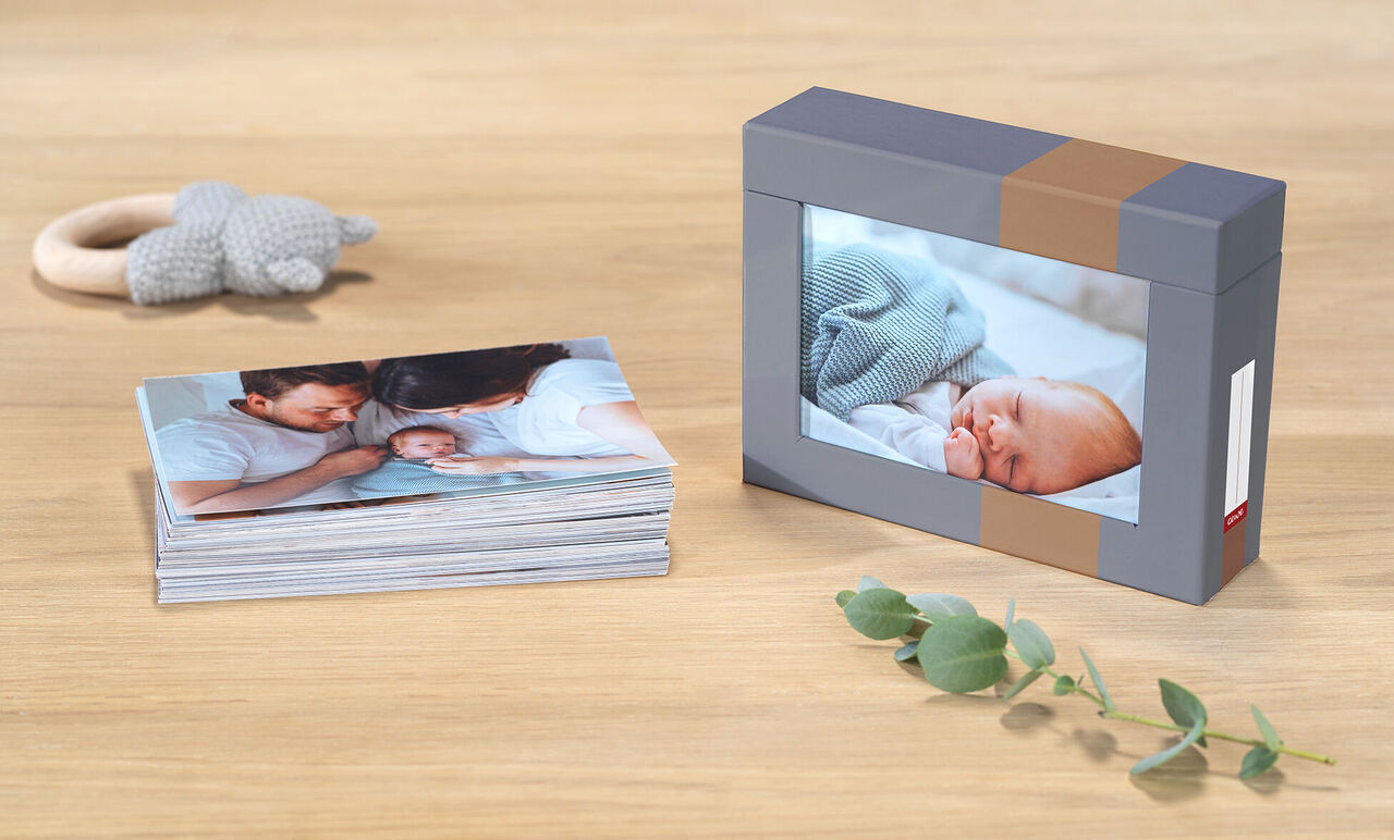 CEWE fotobog med billeder af et nyfødt barn og familie på et træbord, ved siden af en kasse med billede af den sovende baby