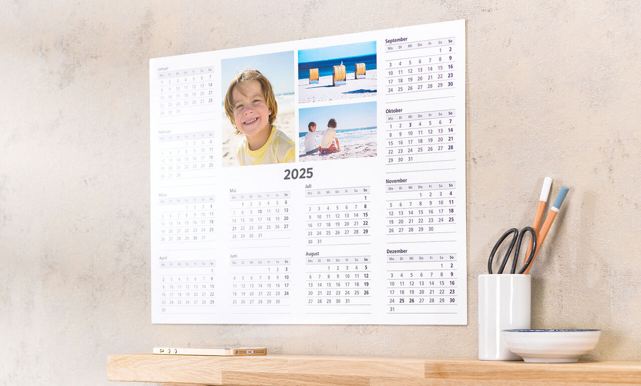 En vægkalender for 2025 med billeder fra strandferier og et tiltalende design. Den hænger over et træbord, hvor en smuk beholder med penne står.
